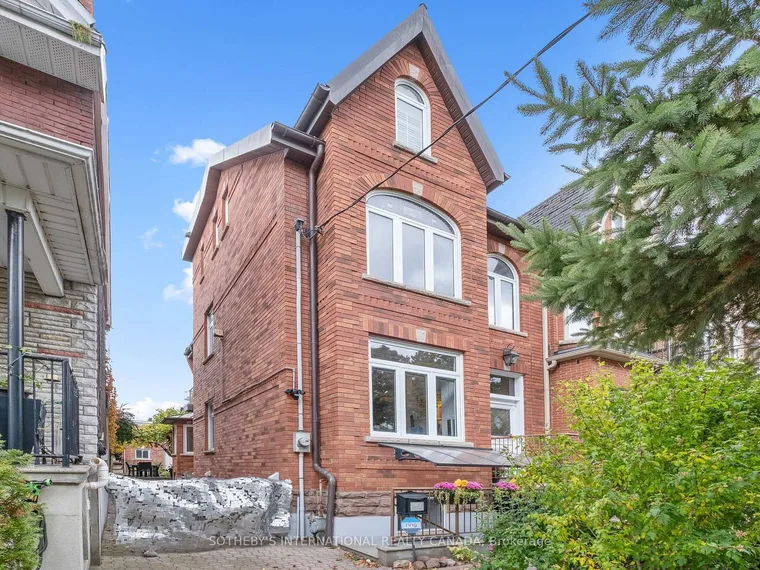 Tall three-storey red-brick Victorian semi with arched windows and a peaked gable behind street trees at 114 Margueretta Street, Toronto, Ontario