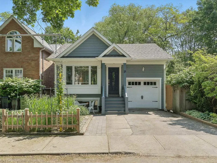 Grey-blue sided storey-and-a-half house with a white garage door, front steps and a wide driveway among leafy gardens at 1A Bowmore Road, Toronto, Ontario